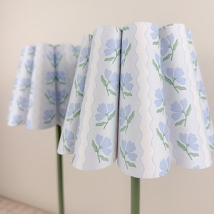 May include: Two lampshades with a floral pattern. The shades are white with a repeating pattern of blue flowers and green leaves. The lampshades have a scalloped edge and are mounted on green stems.