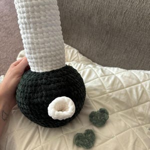 May include: A crocheted plush toy shaped like a bong. The bong is white and green. The toy is sitting on a white blanket. There are two small green crocheted hearts in front of the bong.