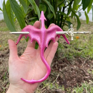 May include: A pink dragon-shaped hair accessory with a silver sword pin. The dragon has outstretched wings and a curled tail. The accessory is held in a hand against a background of green foliage.