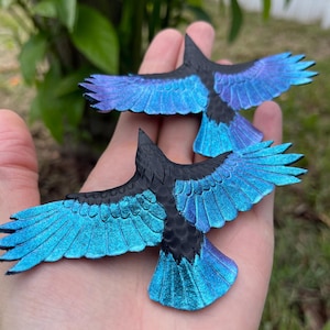 Peut inclure: Deux figurines de corbeaux artisanales aux ailes déployées. Les corbeaux ont des corps noirs et des ailes irisées bleues et violettes. Les figurines sont tenues dans une main sur un fond vert flou.