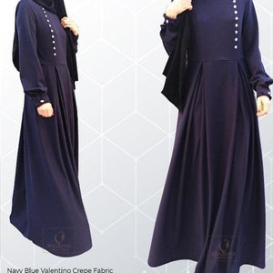 May include: Navy blue maxi dress with white buttons down the left side. The dress has a pleated skirt and is made of a crepe fabric. A black headscarf is included.