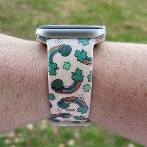 May include: A pink and green watch band with a rainbow and shamrock pattern. The band is made of silicone and is compatible with Apple Watch.