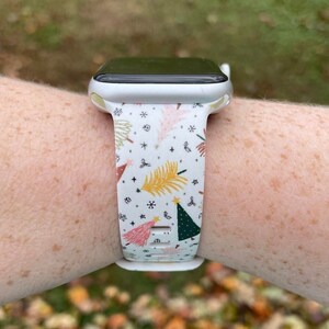 May include: A white Apple Watch with a white band featuring a Christmas tree pattern in pink, green, yellow, and gold.
