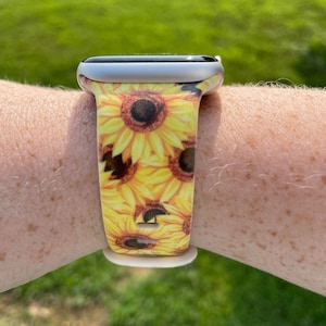 May include: A silver Apple Watch with a sunflower print band. The band is yellow and features a pattern of sunflowers with brown centers.