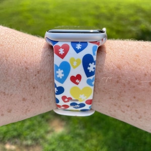 May include: A white Apple Watch band with a pattern of blue, yellow, and red hearts with puzzle pieces in the center.