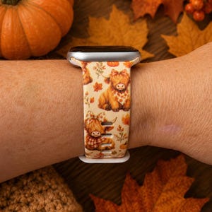 May include: A silicone watch band featuring a cream-colored background with a repeating pattern of brown Highland cows wearing plaid scarves and bows, interspersed with orange pumpkins and autumn leaves, attached to a silver-toned watch casing.