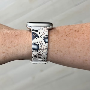 May include: A white Apple Watch band with a black and white print of potion bottles, skulls, stars, and moons. The band is made of silicone and has a silver buckle.