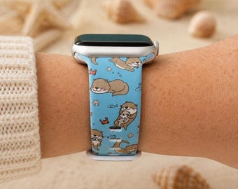 May include: A light blue silicone watch band featuring a repeating pattern of brown otters, small crabs, and seashells. The band is attached to a silver-toned smartwatch case, showcasing a playful aquatic theme with various otter poses.