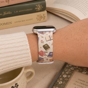 May include: A silicone smartwatch band featuring a cream-colored background with a repeating pattern of various illustrated books in shades of green, blue, pink, and brown, accented with small floral motifs and a silver-toned metal watch casing.