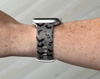 Könnte beinhalten: Eine silberne Apple Watch mit einem schwarzen und grauen Armband mit Fledermaus- und Sternenmuster. Das Armband ist aus Silikon und hat einen sportlichen Look.