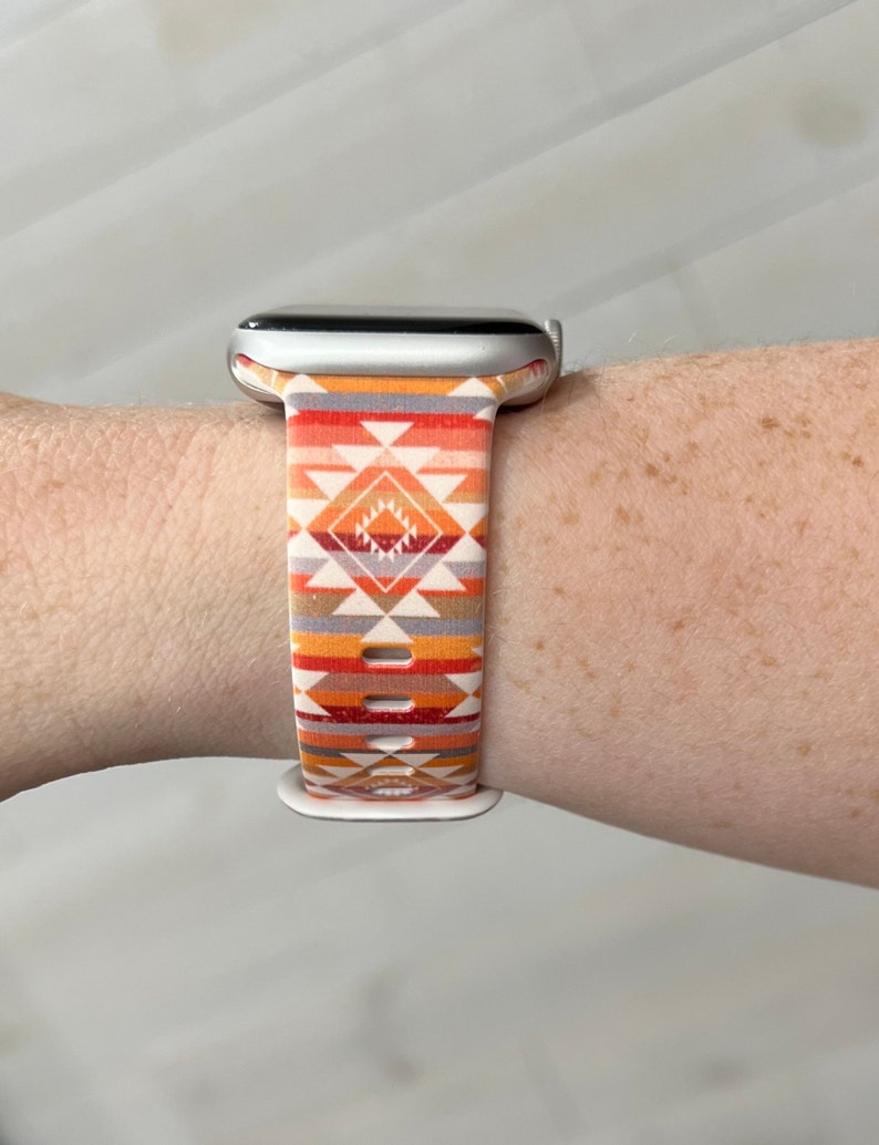 May include: A silver Apple Watch with a white and orange geometric patterned band.