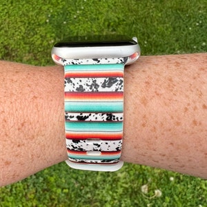 May include: A white Apple Watch band with a colorful southwestern-style pattern. The band has a turquoise, orange, and white striped pattern with black cowhide spots.
