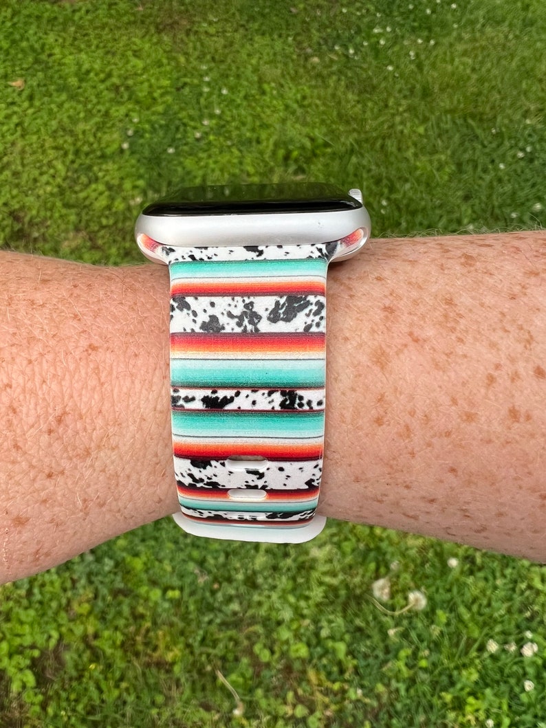 May include: A white Apple Watch band with a colorful southwestern-style pattern. The band has a turquoise, orange, and white striped pattern with black cowhide spots.