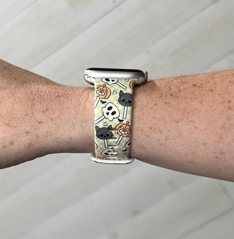 May include: A white Apple Watch band with a green, white and orange Halloween-themed print featuring pumpkins, ghosts and cats.