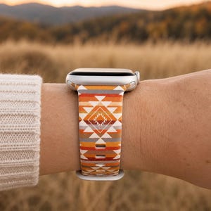 May include: A silver smartwatch featuring a silicone band with a geometric pattern in shades of orange, white, and grey. The band displays a repeating diamond motif with horizontal stripes, secured with a standard buckle closure on the wrist.