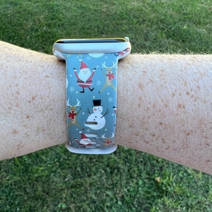 May include: A white Apple Watch with a blue and white Christmas-themed band. The band features Santa Claus, reindeer, and a snowman.