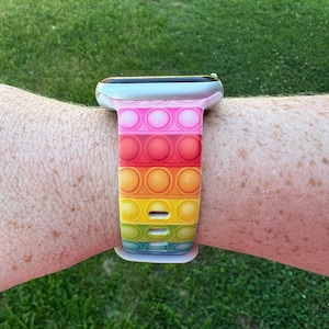 May include: A rainbow-colored silicone watch band with a pop-it design. The band is on a silver smartwatch.