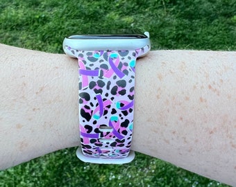 May include: A white Apple Watch with a pink, black, and purple leopard print band featuring purple ribbon designs.