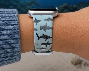 Puede incluir: Una correa de reloj de silicona azul claro con un patrón repetitivo de varias siluetas de tiburones gris oscuro, incluyendo tiburones martillo y grandes tiburones blancos, unida a una caja de reloj inteligente plateada con acabado mate.