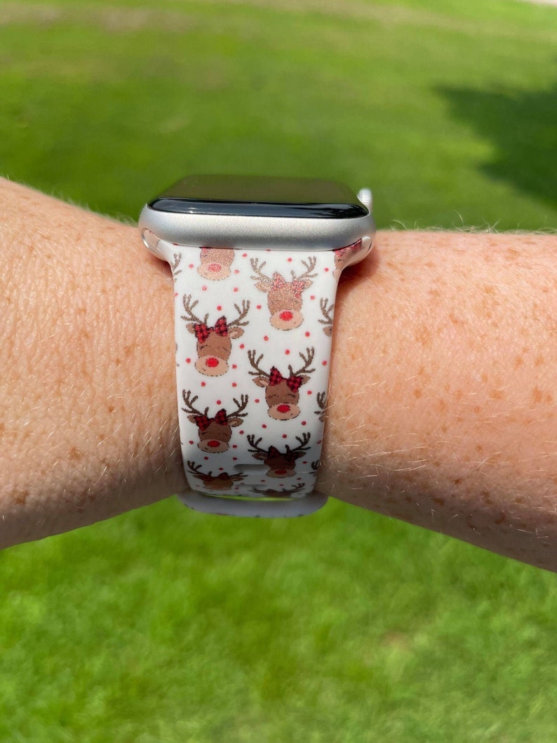 May include: A white Apple Watch with a white and red reindeer patterned band. The band has red bows and white polka dots.