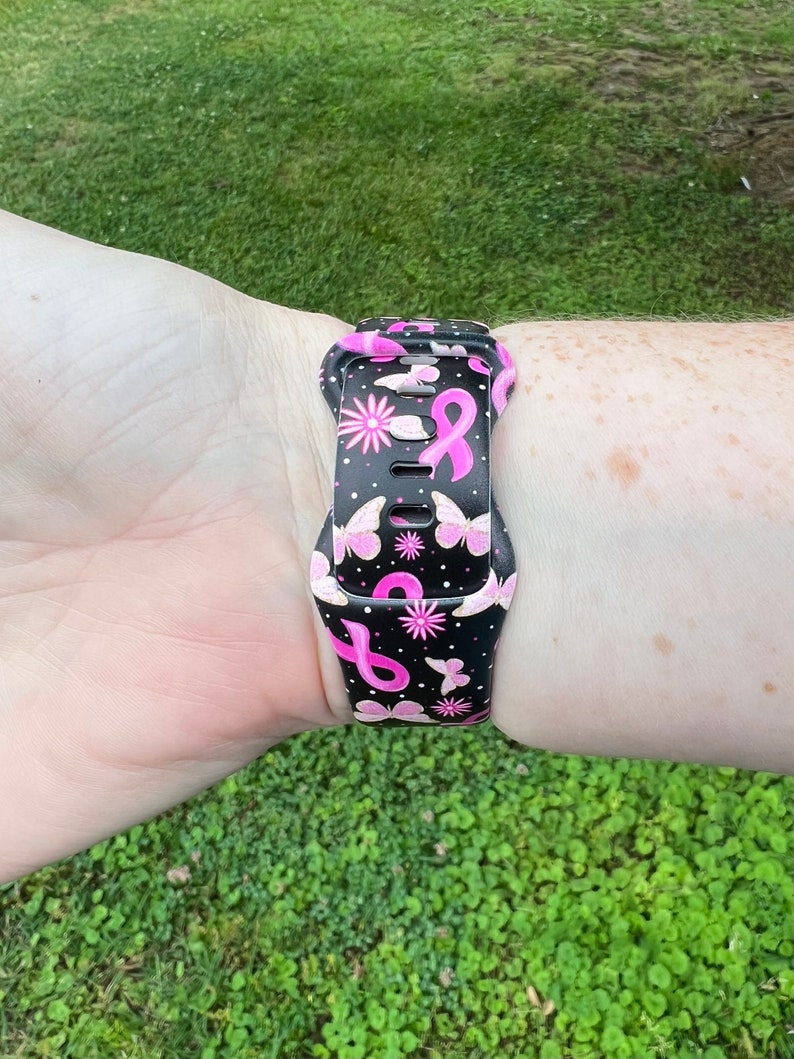 May include: A black watch strap with a pink ribbon and butterfly pattern. The strap is made of silicone and is compatible with most smartwatches.