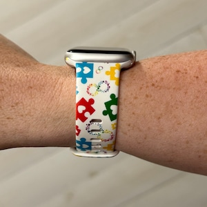 May include: White Apple Watch band with a colorful puzzle piece and heart design. The band is made of silicone and has a white background with multi-colored puzzle pieces and hearts.