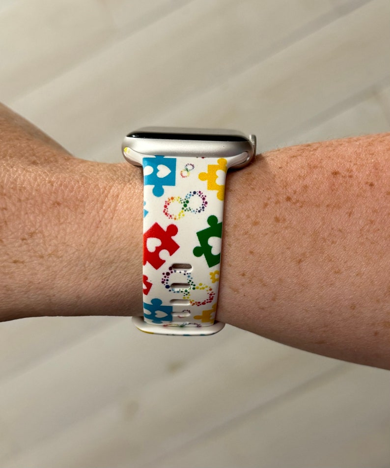 May include: White Apple Watch band with a colorful puzzle piece and heart design. The band is made of silicone and has a white background with multi-colored puzzle pieces and hearts.