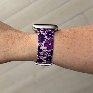 May include: A purple and white floral patterned Apple Watch band. The band is made of silicone and has a silver buckle.