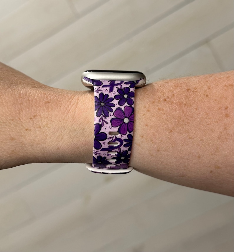 May include: A purple and white floral patterned Apple Watch band. The band is made of silicone and has a silver buckle.