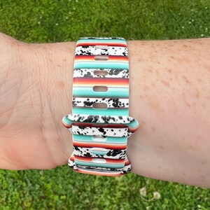 May include: A colorful watch band with a southwestern pattern. The band is white with black, red, and turquoise stripes. It also has a cowhide print.