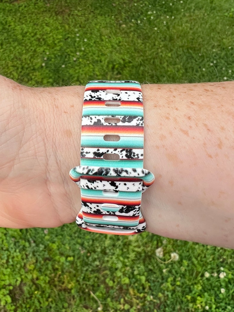 May include: A colorful watch band with a southwestern pattern. The band is white with black, red, and turquoise stripes. It also has a cowhide print.