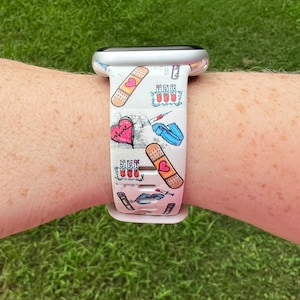 May include: White Apple Watch band with a medical theme. The band features illustrations of bandages, hearts, syringes, and test tubes. The design is in shades of red, pink, blue, and white. The watch band is on a wrist with green grass in the background.