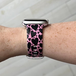 May include: A pink and black patterned watch band with a heart design. The band is made of silicone and is compatible with Apple Watch.
