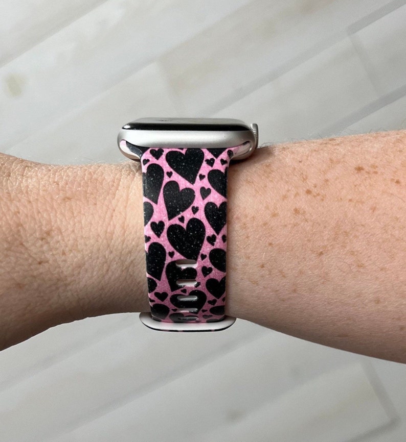 May include: A pink and black patterned watch band with a heart design. The band is made of silicone and is compatible with Apple Watch.