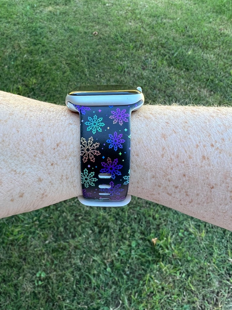 May include: A black smartwatch band with a colorful snowflake pattern. The band is made of silicone and has a white buckle.