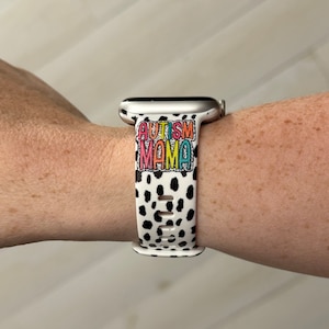 May include: White and black spotted Apple Watch band with the words "AUTISM MAMA" in colorful letters. The band is made of a flexible material and has multiple holes for adjustment.
