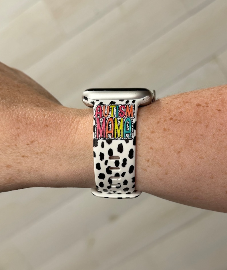 May include: White and black spotted Apple Watch band with the words "AUTISM MAMA" in colorful letters. The band is made of a flexible material and has multiple holes for adjustment.