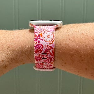 May include: A white smartwatch with a pink and red patterned band. The band features a repeating design of various shapes and floral elements. The watch is secured on a wrist.