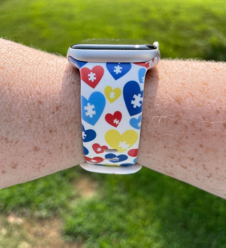 May include: A white Apple Watch band with a pattern of blue, yellow, and red hearts with puzzle pieces in the center.