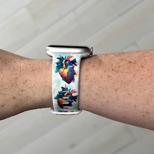 May include: A white Apple Watch band with a colorful anatomical heart print. The band is made of silicone and has a silver buckle.