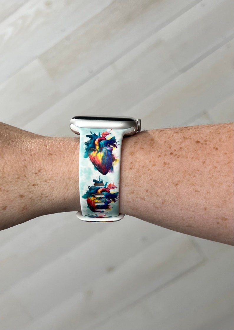 May include: A white Apple Watch band with a colorful anatomical heart print. The band is made of silicone and has a silver buckle.