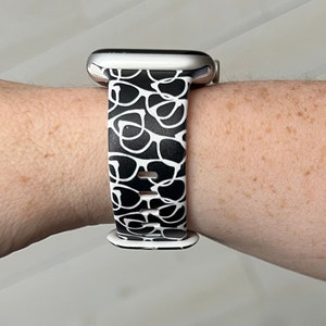 May include: A silver Apple Watch with a black and white patterned band. The band has a design of white eyeglasses on a black background.