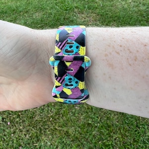 May include: A colorful watch band with a patchwork design featuring black, yellow, pink, and blue colors. The band is made of silicone and has a buckle closure.