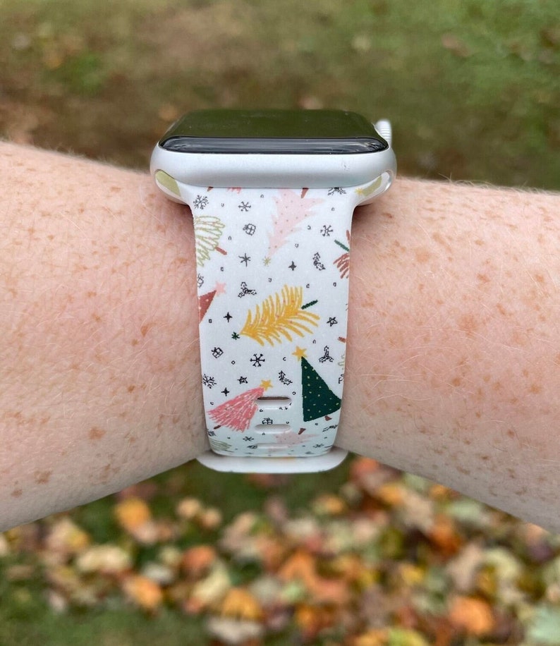 May include: A white Apple Watch with a white band featuring a Christmas tree pattern in pink, green, yellow, and gold.
