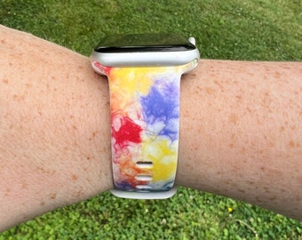 May include: A white Apple Watch with a colorful tie-dye patterned band. The band has shades of yellow, red, blue, and white.