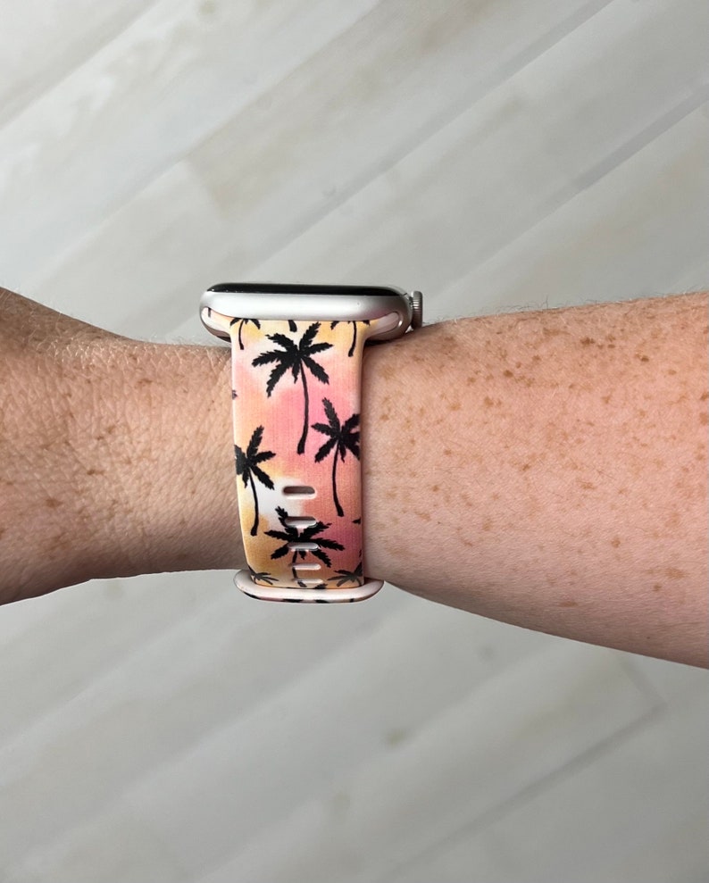 K&ouml;nnte beinhalten: Eine silberne Smartwatch mit einem pinkfarbenen und orangen Armband mit Palmenmuster.