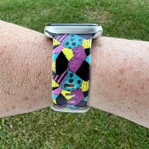 May include: A black, white, yellow, and purple patterned watch band with a silver watch face.