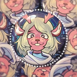 May include: A close-up of a round button featuring a cartoon character with pink skin, white fur, and antlers. The character has large, green eyes and a pink nose. The button has a white border with a dotted pattern.