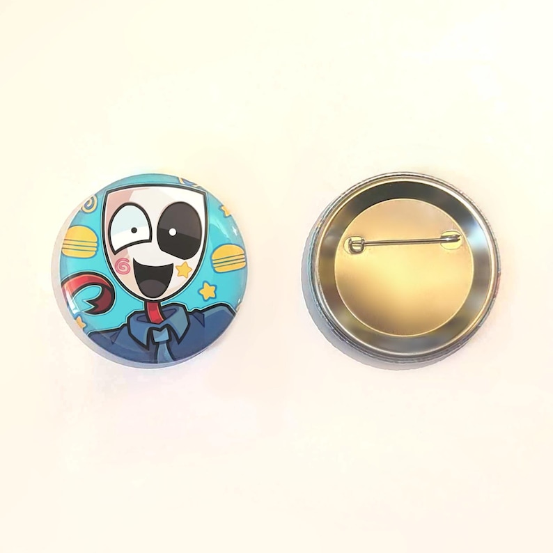Op de afbeelding: Twee ronde pins. E&eacute;n pin toont een stripfiguur met een wit masker, een blauw overhemd en een rode stropdas. De achtergrond is lichtblauw met gele sterren en hamburger illustraties. De andere pin is zilverkleurig.