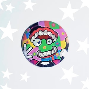 May include: A colorful round button with a cartoon character with a large mouth, green eyes, and a red hat. The character is on a background of colorful squares and stars.